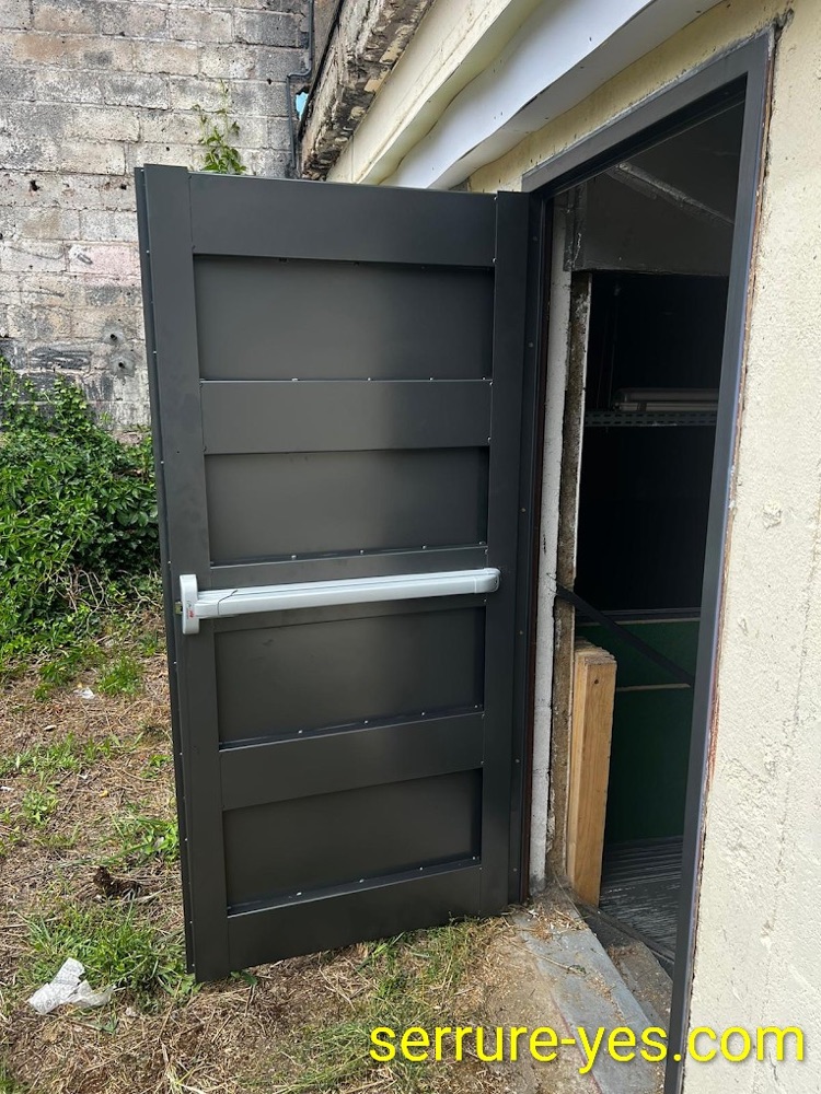 Porte métallique noire ouverte, équipée d'une barre anti-panique horizontale, donnant sur un local de stockage