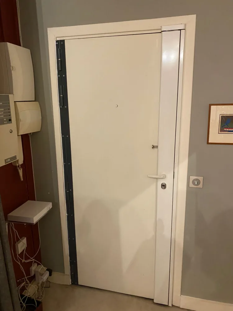 Porte blindée blanche avec barre de pivot anthracite visible côté paumelles et carénage blanc côté serrure